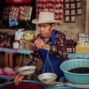 Elderly man in colorful attire weighing beans at a vibrant market stall.