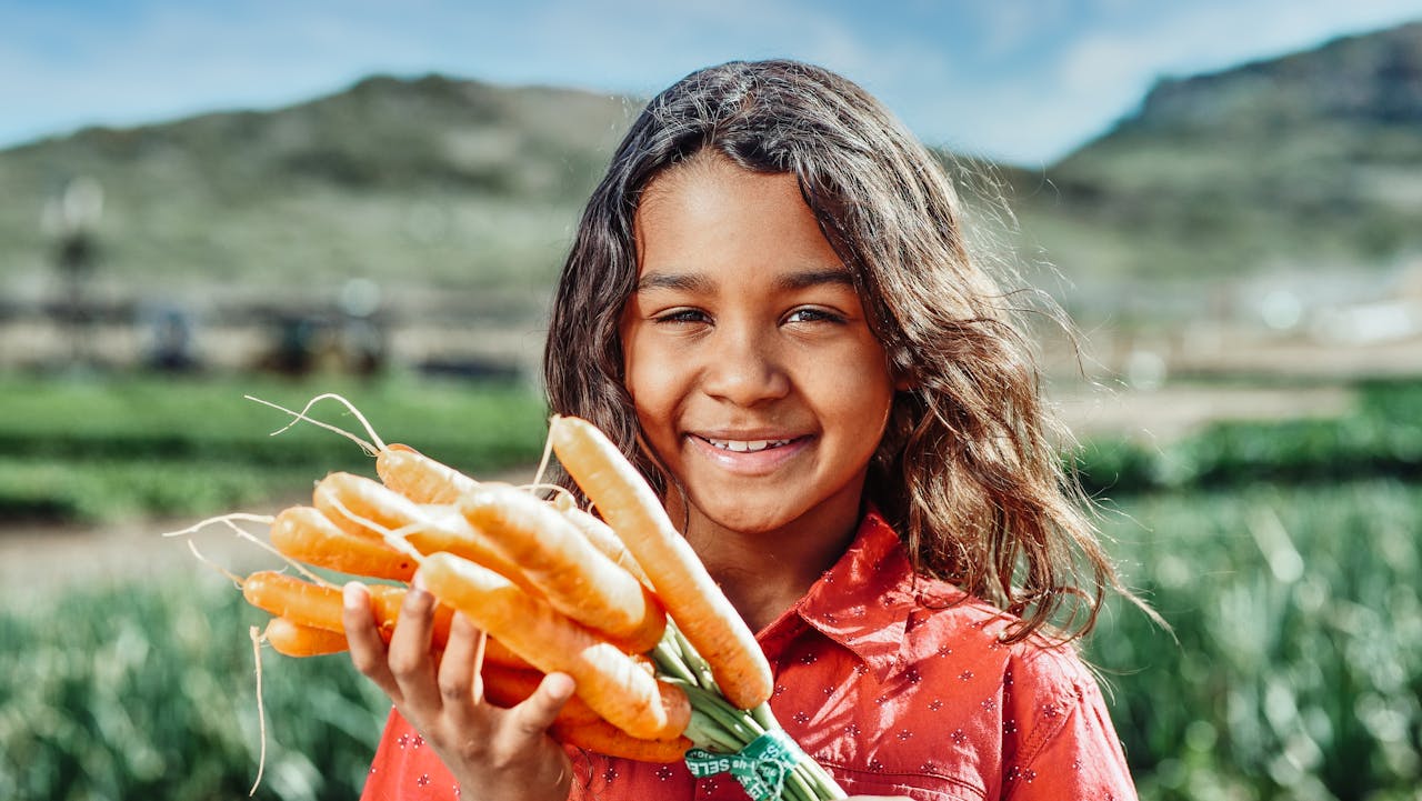 A joyful young girl holding fresh organic carrots in a sunny countryside farm setting.